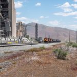 Trem de carga passando diante de uma grande instalação industrial, com montanhas ao fundo.