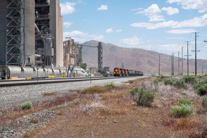 Trem de carga passando diante de uma grande instalação industrial, com montanhas ao fundo.