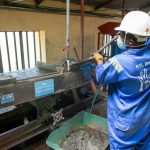 Trabalhador de macacão azul opera uma mesa vibratória para processamento de minério em uma instalação industrial.