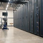 Fileiras de gabinetes metálicos com servidores, dispositivos de rede e cabos em um data center moderno.