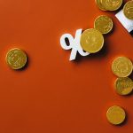 Moedas douradas espalhadas sobre um gráfico financeiro, acompanhadas de um símbolo de porcentagem em fundo laranja.