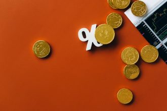 Moedas douradas espalhadas sobre um gráfico financeiro, acompanhadas de um símbolo de porcentagem em fundo laranja.
