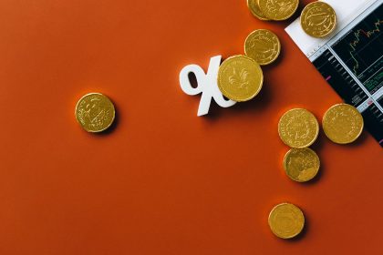 Moedas douradas espalhadas sobre um gráfico financeiro, acompanhadas de um símbolo de porcentagem em fundo laranja.