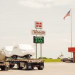 Posto de combustível rural nos Estados Unidos com bomba de diesel, caminhões estacionados e bandeira americana.