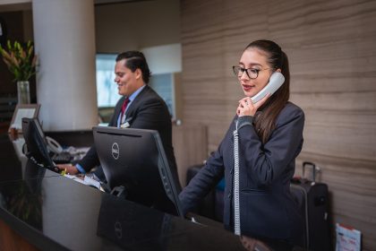 Dois profissionais trabalham no balcão de recepção de um hotel, atendendo uma chamada telefônica.